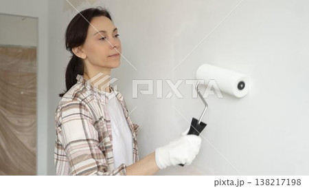 Middle aged woman wearing gloves is skillfully applying paint to a white wall with a paint roller in an apartment undergoing renovation, transforming the space with her creative touch Middle aged woman wearing gloves is skillfully applying paint to a white wall with a paint roller in an apartment undergoing renovation, transforming the space with her creative touch 138217198