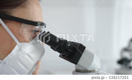Female scientist peering into microscope, analyzing biological sample with precision in sterile laboratory research environment. Medicine and science concept 138217201