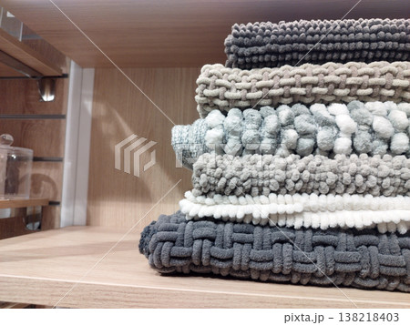 A stack of cozy, textured knit blankets in soothing shades of grey, beige, and white rests on a clean wooden shelf. The varied patterns, including chunky weaves and pom-poms, evoke a warm, homey feel 138218403