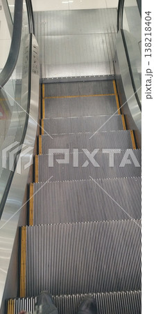 A high-angle, first-person perspective looking down a modern escalator in a bright, clean building. The metallic steps with yellow safety lines lead the eye toward the landing, reflecting a minimalist A high-angle, first-person perspective looking down a modern escalator in a bright, clean building. The metallic steps with yellow safety lines lead the eye toward the landing, reflecting a minimalist 138218404