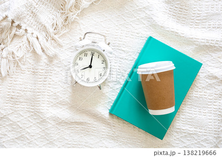 White alarm clock, paper disposable cup and green book in white bed, top view. 138219666