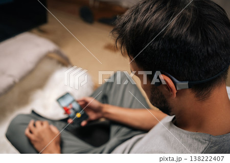 Man wearing bone conduction headphones, relaxing indoors and listening to audio from smartphone, enjoying modern technology for immersive sound experience and personal entertainment time. Man wearing bone conduction headphones, relaxing indoors and listening to audio from smartphone, enjoying modern technology for immersive sound experience and personal entertainment time. 138222407