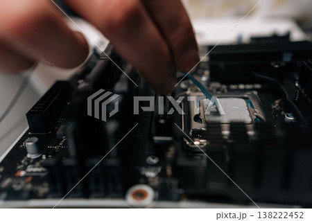 Close-up hands of unrecognizable technician applying thermal paste on computer processor, spreading precise layer for efficient cooling and improved performance during hardware maintenance. 138222452