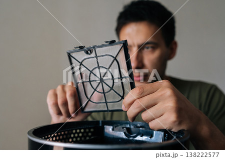 Headshot of focused young man checking air humidifier filter, carefully examining device component for maintaining indoor air quality and home wellness. Concept of maintenance modern home appliance. Headshot of focused young man checking air humidifier filter, carefully examining device component for maintaining indoor air quality and home wellness. Concept of maintenance modern home appliance. 138222577
