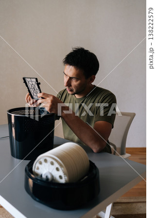 Vertical portrait of focused man holding inspecting air humidifier filter, performing maintenance and cleaning to ensure clean, healthy air in home environment. Concept of maintenance home appliance. Vertical portrait of focused man holding inspecting air humidifier filter, performing maintenance and cleaning to ensure clean, healthy air in home environment. Concept of maintenance home appliance. 138222579