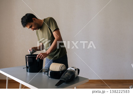 Portrait of young man disassembling and cleaning air humidifier filter standing by table in living room, performing regular maintenance for healthy air quality. Concept of household cleanliness. 138222583