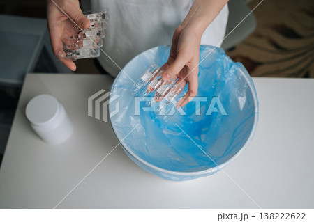 Top view of person hands dropping multiple glass ampoules of expired medication into blue plastic garbage bag inside white trash can, illustrating proper and safe disposal of pharmaceutical waste. 138222622