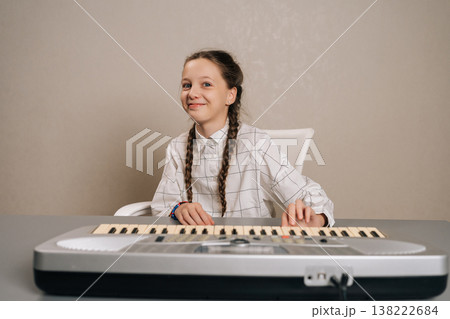 Studio portrait of happy young girl with pigtails smiling looking at camera and sitting at table, playing digital keyboard synthesizer, engaging in musical practice and expressing creativity. 138222684