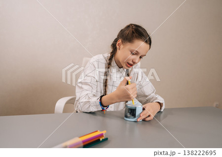 Focused young girl with pigtails sharpening pencil in electric sharpener, sitting at desk with colorful drawing tools, focusing on preparing supplies for learning and artwork. 138222695