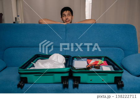 Dreaming traveler male planning or preparing for upcoming journey, looking thoughtfully at open suitcase full of packed clothes and travel essentials on blue sofa in cozy home setting. Dreaming traveler male planning or preparing for upcoming journey, looking thoughtfully at open suitcase full of packed clothes and travel essentials on blue sofa in cozy home setting. 138222711