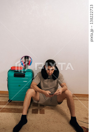 Male traveler sitting on floor with neck pillow and smartphone, having a delay, waiting for departure, feeling impatient while checking updates on mobile device during trip. Male traveler sitting on floor with neck pillow and smartphone, having a delay, waiting for departure, feeling impatient while checking updates on mobile device during trip. 138222713