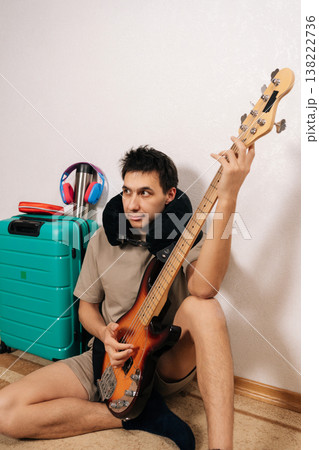 Portrait of young man in travel neck pillow sits by a turquoise suitcase in an airport terminal, playing bass guitar and wearing headphones relaxed, ready for a musical journey and travel adventure. Portrait of young man in travel neck pillow sits by a turquoise suitcase in an airport terminal, playing bass guitar and wearing headphones relaxed, ready for a musical journey and travel adventure. 138222736