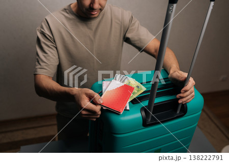 Close-up hand of traveler organizing essential documents and foreign currency into a turquoise suitcase, ensuring all travel preparations are complete for an upcoming trip or international vacation. 138222791