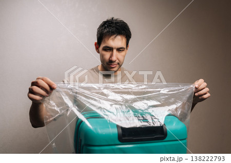 Portrait of focused traveler male wraps hard-shell suitcase in clear protective plastic on a table, preparing luggage for travel and ensuring belongings are secure and ready for departure. Portrait of focused traveler male wraps hard-shell suitcase in clear protective plastic on a table, preparing luggage for travel and ensuring belongings are secure and ready for departure. 138222793