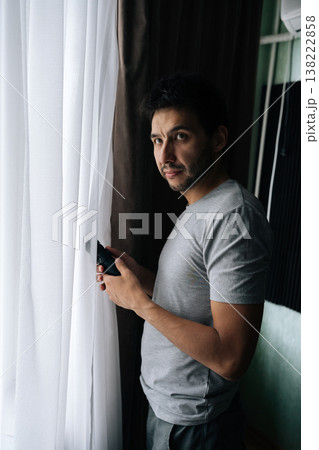 Vertical portrait of young bearded man spying with monocular, peeking through window curtains, observing outside, engaging in surveillance, curiosity and undercover investigation. Vertical portrait of young bearded man spying with monocular, peeking through window curtains, observing outside, engaging in surveillance, curiosity and undercover investigation. 138222858