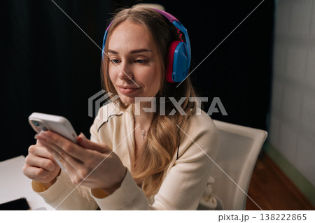 Attractive blonde young woman enjoying leisure time at home in evening, wearing vibrant headphones and engaging with mobile phone, finding entertainment and communication in dark relaxed setting. Attractive blonde young woman enjoying leisure time at home in evening, wearing vibrant headphones and engaging with mobile phone, finding entertainment and communication in dark relaxed setting. 138222865