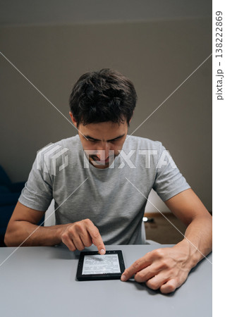 Vertical portrait of focused young adult man using touchscreen e-reader siting at home desk, scrolling through e-book with finger, focused on portable device for study, leisure reading or research. 138222869