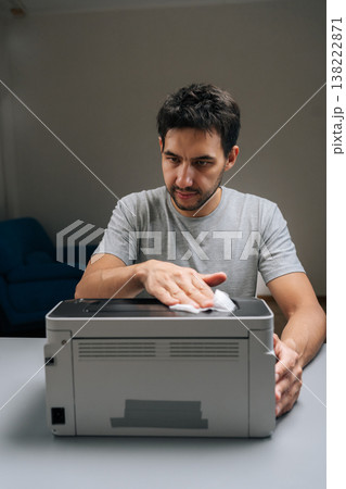 Vertical portrait of man performing printer maintenance, wiping dust off office laser printer, ensuring technology hygiene, cleaning and servicing electronic equipment in home office environment. 138222871