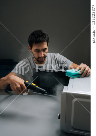Portrait of man cleaning microwave oven with steam cleaner and sponge, using hot vapor for powerful disinfection and removing dirt, representing effective household chore and modern cleaning solution. 138222877