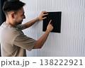 Smiling man installing black acoustic foam panel on white decorative wall to reduce echo, improve sound quality in a home studio or recording space during renovation project. 138222921