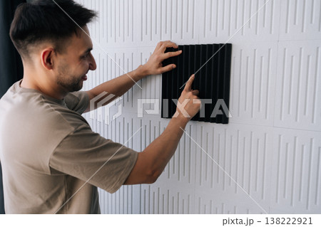 Smiling man installing black acoustic foam panel on white decorative wall to reduce echo, improve sound quality in a home studio or recording space during renovation project. 138222921