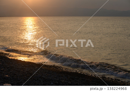 Sunset casts golden reflections over serene waters and pebbled shoreline in a tranquil coastal setting Sunset casts golden reflections over serene waters and pebbled shoreline in a tranquil coastal setting 138224968