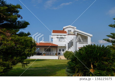 Elegant white villa surrounded by lush greenery under a bright blue sky in a serene landscape Elegant white villa surrounded by lush greenery under a bright blue sky in a serene landscape 138225075