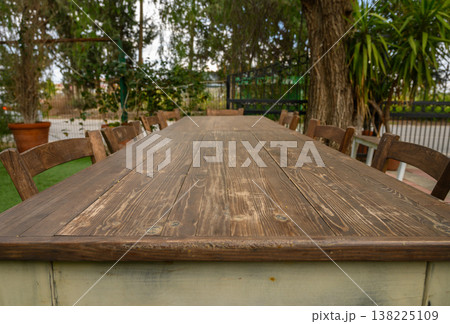 Long wooden table set for gathering in a peaceful outdoor garden during a sunny Long wooden table set for gathering in a peaceful outdoor garden during a sunny 138225109