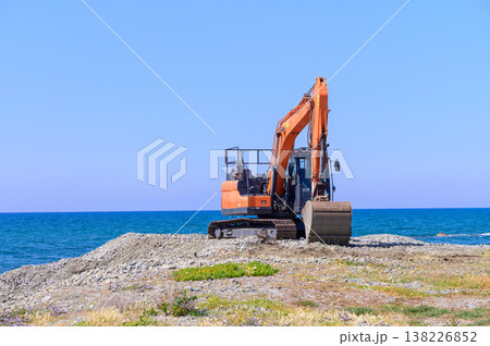 An orange excavator stands idle on a coastal site in Cyprus, framed by the blue waters of the Mediterranean and rugged shoreline 138226852
