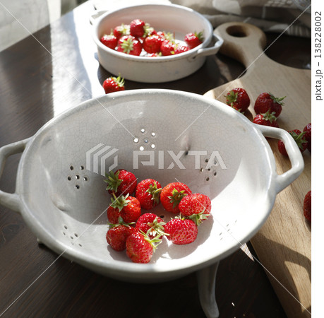 Fresh organic strawberries in white crackle glazed ceramic colander. High contrast photography of red berries on dark wood table with cinematic sunlight shadows and wabi-sabi aesthetic. 138228002