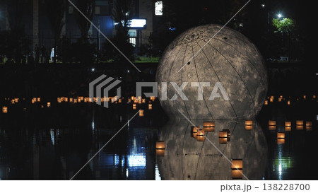 Inflatable moon sculpture floating on a dark river with many illuminated paper lanterns at night. 138228700
