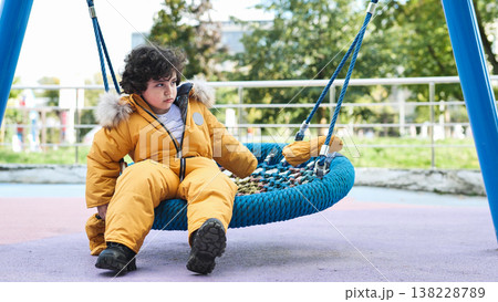 Thoughtful boy wearing warm yellow winter jumpsuit sitting on playground swing Thoughtful boy wearing warm yellow winter jumpsuit sitting on playground swing 138228789