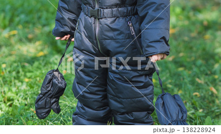 Child holding mittens, wearing winter snowsuit, standing outdoors in green grass Child holding mittens, wearing winter snowsuit, standing outdoors in green grass 138228924