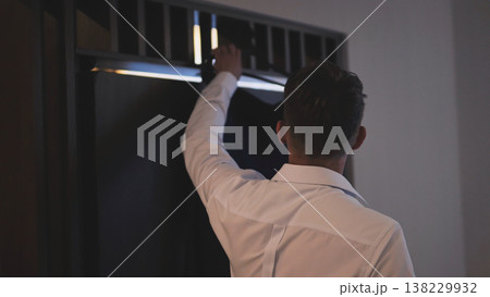 Rear view of a groom in a white dress shirt getting his suit jacket from a dark wardrobe. Rear view of a groom in a white dress shirt getting his suit jacket from a dark wardrobe. 138229932