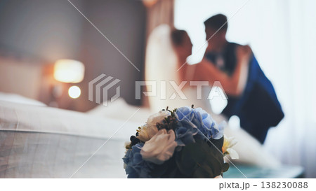 Blue and white wedding bouquet in the foreground with a blurred just married couple embracing in the background in their hotel room Blue and white wedding bouquet in the foreground with a blurred just married couple embracing in the background in their hotel room 138230088