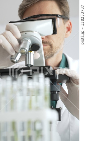 Man scientific researcher in protective gear analyzing specimens through a microscope in a sterile laboratory environment. Medicine, health care and science concept Man scientific researcher in protective gear analyzing specimens through a microscope in a sterile laboratory environment. Medicine, health care and science concept 138233737