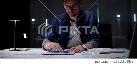 Professional man wearing glasses researching finances on digital tablet, analyzing business data and graphs, working late night in modern office 138233868