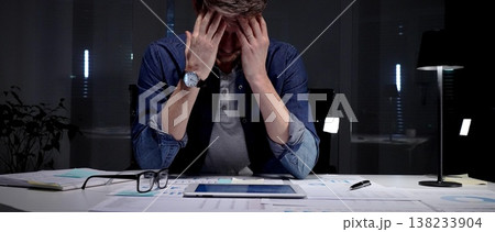 Young businessman with glasses sits at a dark office desk late at night, overwhelmed by documents and a tablet, clutching his head from stress, burnout and exhaustion Young businessman with glasses sits at a dark office desk late at night, overwhelmed by documents and a tablet, clutching his head from stress, burnout and exhaustion 138233904