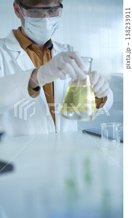 Male scientist wearing white protective gloves, mask and glasses is holding a yellow chemical solution inside an Erlenmeyer flask in a laboratory, vertical portrait. Science and medicine 138233911