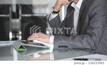 Businessman in a modern office setup, focusing intently on a tablet screen while engaging in strategic thinking, analyzing data, and preparing for an important corporate meeting or presentation 138233983