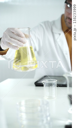 Man scientist wearing white protective gloves, and glasses is holding a yellow chemical solution inside an Erlenmeyer flask in a laboratory, vertical portrait 138233999
