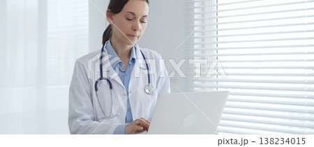 Female doctor working on laptop in white lab coat and stethoscope, sitting at desk in bright, sunlit medical office. Medicine and healthcare concept Female doctor working on laptop in white lab coat and stethoscope, sitting at desk in bright, sunlit medical office. Medicine and healthcare concept 138234015