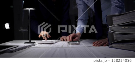Business professionals collaborating over financial reports and a calculator beneath a desk lamp, reviewing documents and data late into the night to plan budget and strategy Business professionals collaborating over financial reports and a calculator beneath a desk lamp, reviewing documents and data late into the night to plan budget and strategy 138234056
