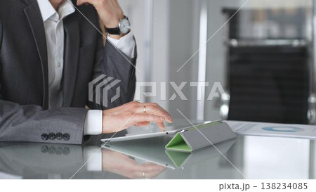 Business professional in a suit and wristwatch using a tablet on a glass desk, focused on data analysis and strategy planning in a modern corporate office environment 138234085