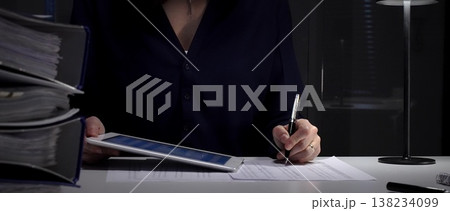 Woman working late, signing documents and analyzing data on a tablet by office lamp light at night. Audit and taxes in business 138234099