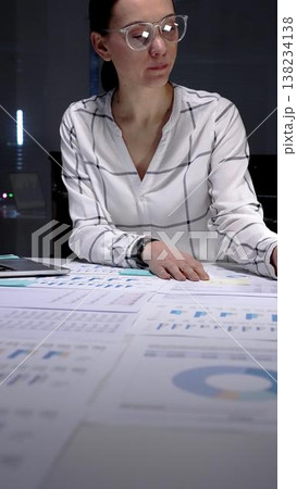 Businesswoman working late under a desk lamp, reviewing charts and financial documents with focused concentration, preparing reports and planning strategy for company growth Businesswoman working late under a desk lamp, reviewing charts and financial documents with focused concentration, preparing reports and planning strategy for company growth 138234138