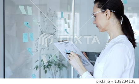 Professional business woman working on successful project strategy, analyzing data on clipboard, developing new ideas on glass whiteboard with sticky notes in modern office Professional business woman working on successful project strategy, analyzing data on clipboard, developing new ideas on glass whiteboard with sticky notes in modern office 138234139