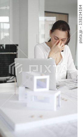 Female architect in eyeglasses concentrating on a detailed building model and blueprints, carefully planning a modern architectural design in a professional office Female architect in eyeglasses concentrating on a detailed building model and blueprints, carefully planning a modern architectural design in a professional office 138234164