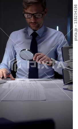 Financial auditor or accountant scrutinizing documents with a magnifying glass and calculator in a dark office. Audit and taxes in business Financial auditor or accountant scrutinizing documents with a magnifying glass and calculator in a dark office. Audit and taxes in business 138234198