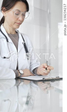 Female doctor in white coat and glasses writing medical notes on a clipboard at clinic desk, managing patient records. Medicine and health care 138234217